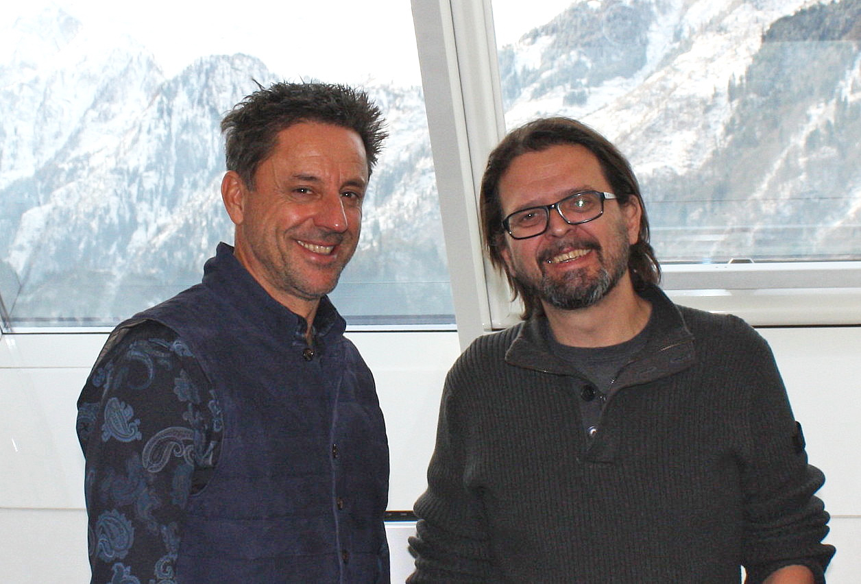 Porträtaufnahmen von Christoph Bründl und Christian Holzer. Stehend vor einem Bergbild. Beide lächeln in die Kamera.