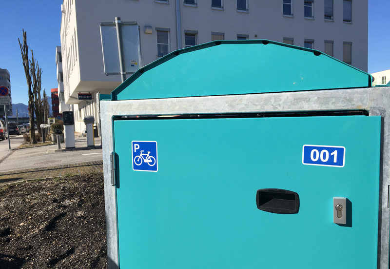 Türkise Radbox im Vordergrund mit zwei blauem Fahrradsymbol, im Hintergrund, Jakob-Haringer-Straße und Bürogebäude Techno 4